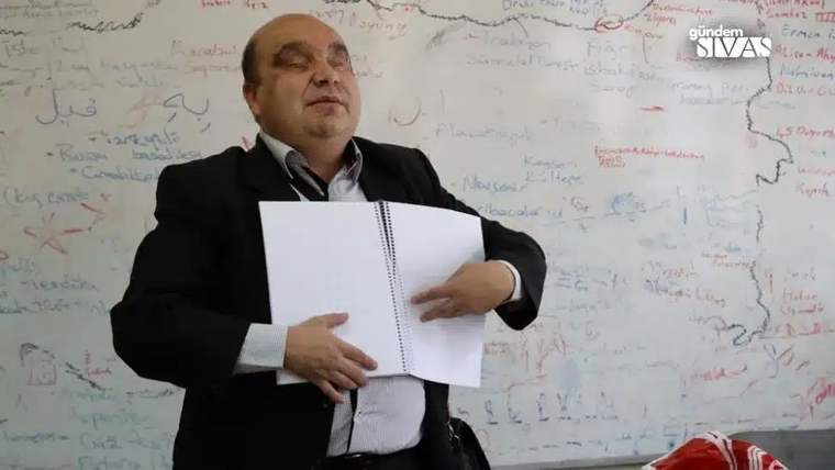 Bir kişi, elinde not defteriyle sınıf ortamında konuşuyor.