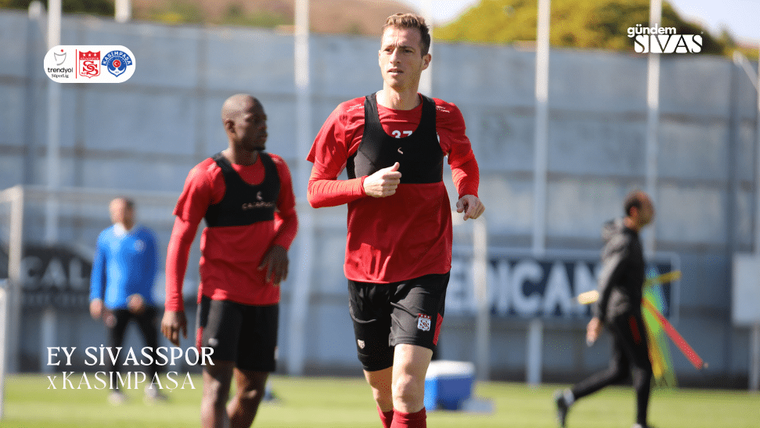 Yeşil sahada kırmızı antrenman formaları giyen iki futbolcu koşuyor; öndeki oyuncu kadrajın merkezinde.