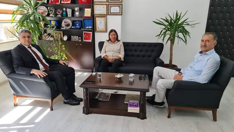 Üç kişi, bir ofiste oturmuş, masanın etrafında sohbet ediyor. Arkada çeşitli ödüller ve belgeler sergileniyor.