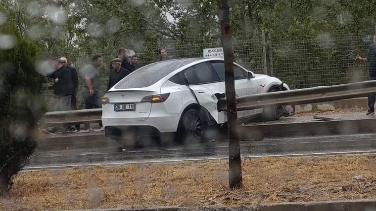 Beyaz Tesla, yağmur altında yol kenarındaki bariyere dayanmış; çevresinde birkaç kişi duruyor.
