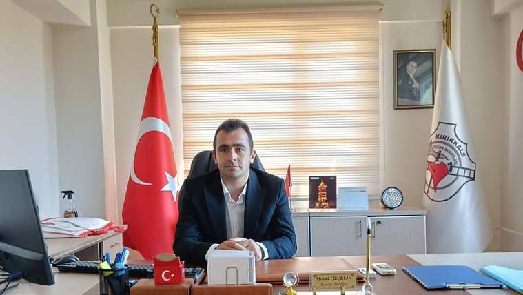 Masa başında oturan bir kişi, Türk bayrağı ve itfaiye amblemli bayrağın önünde kameraya bakıyor.