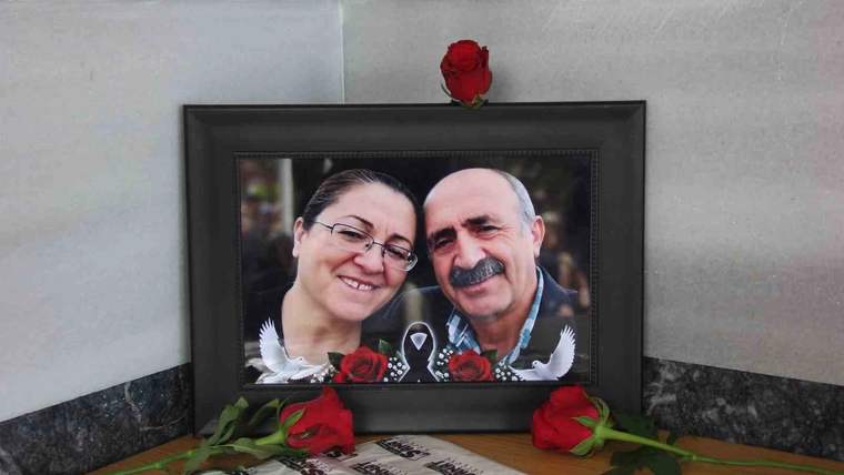 Siyah çerçeveli portre fotoğrafının önünde kırmızı güller ve üzerinde yazılar bulunan örtü yer alıyor.