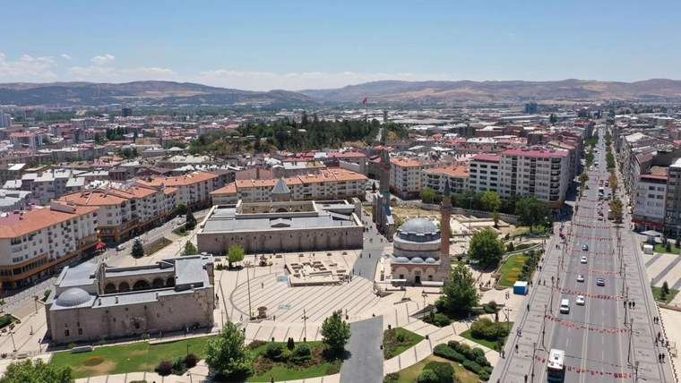 Havadan görülen kent merkezinde cami kompleksi, apartmanlar, ağaçlı yollar ve ilerleyen araçlar yer alıyor.