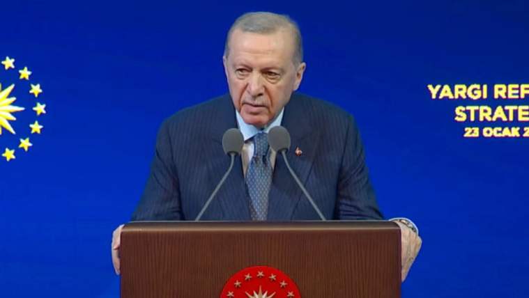 Kürsüde konuşan bir kişi, arkasında mavi bir fon ve yazılarla birlikte duruyor.