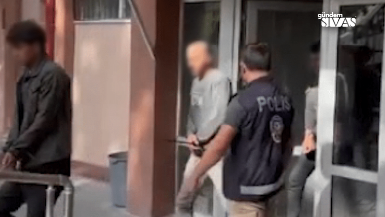 Üzerinde POLİS yazan görevli, elleri önde bir kişiyle bina girişinde ilerliyor; başka biri kapıda bekliyor.