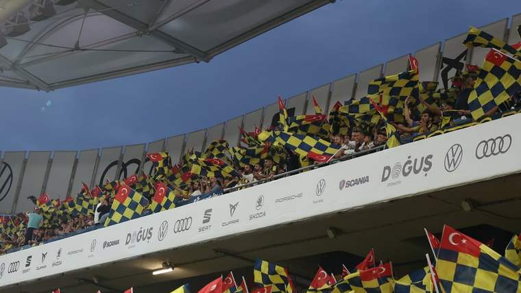 Stadyum tribünlerinde sarı-lacivert damalı bayraklar ve Türk bayrakları taşıyan taraftarlar görülüyor.