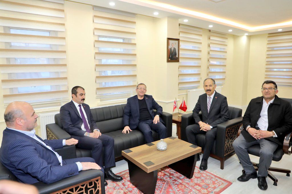 Bir grup erkek, bir ofis ortamında oturuyor; masada bir masa lambası ve bir vazo var.