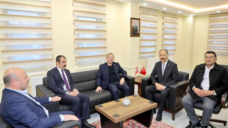 Bir grup erkek, bir ofis ortamında oturuyor; masada bir masa lambası ve bir vazo var.