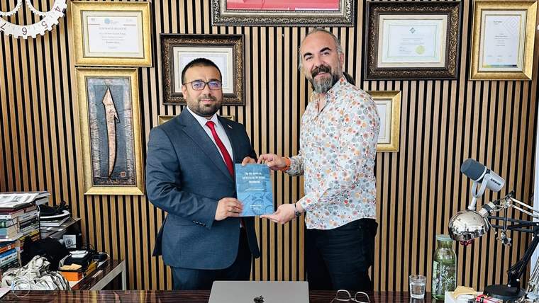 İki kişi, ofis ortamında bir belgeyi birbirine uzatıyor. Arka planda çerçevelenmiş belgeler ve Türk bayrağı var.