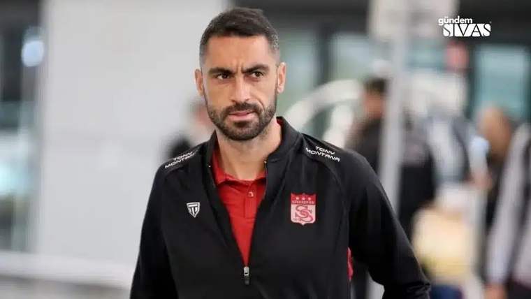 Koyu renkli bir antrenman kıyafeti giymiş bir futbolcu, ciddi bir ifadeyle yürürken görünüyor.