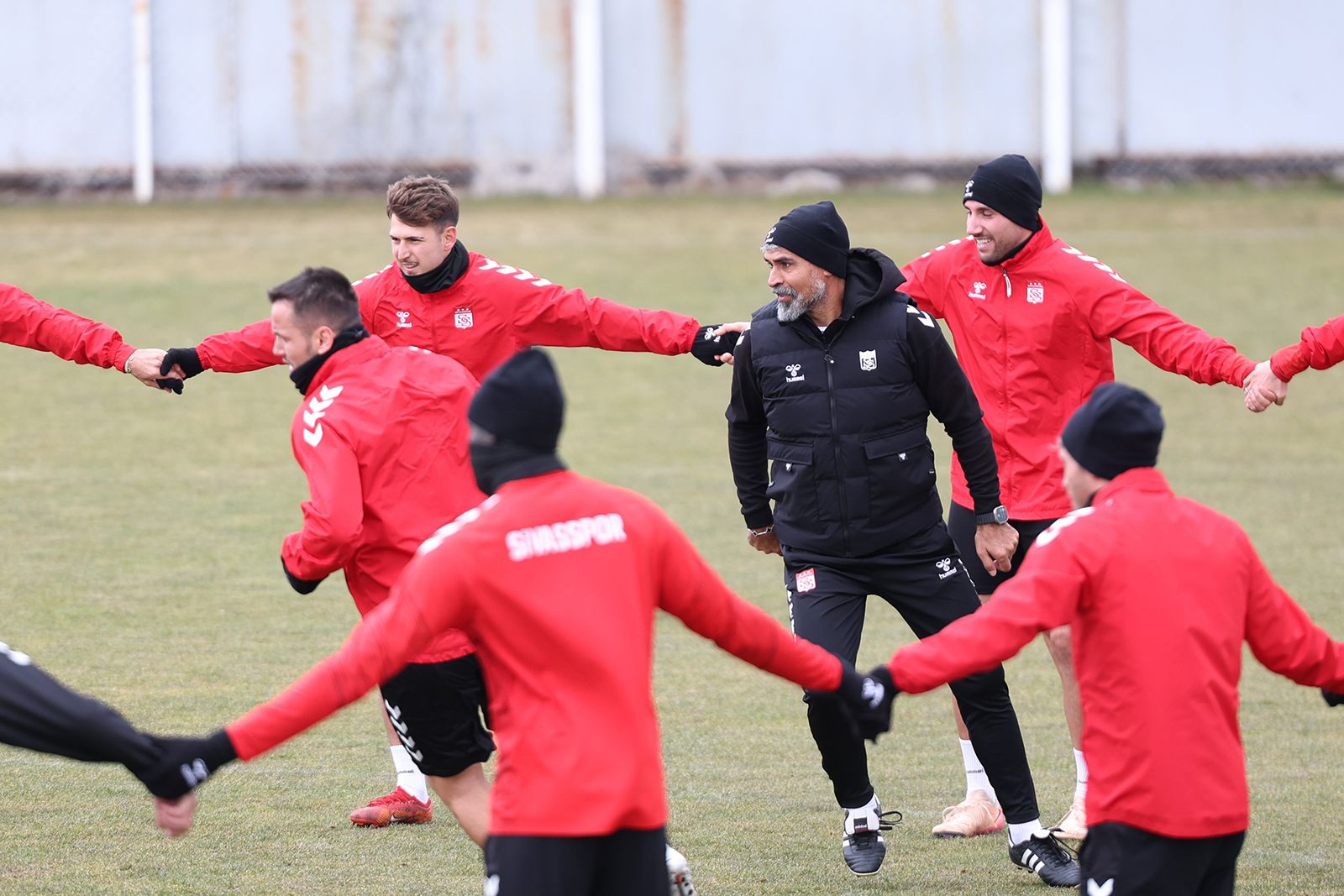 Sivasspor yazılı kırmızı eşofmanlı futbolcular, sahada el ele tutuşarak ısınırken ortalarında siyah giyimli bir kişi duruyor.