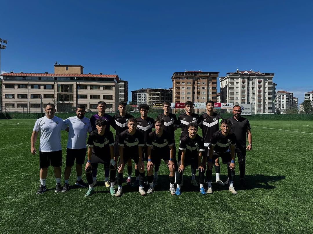 Futbol sahasında siyah formalı genç oyuncular ve iki kişi, şehir binaları önünde takım halinde duruyor.