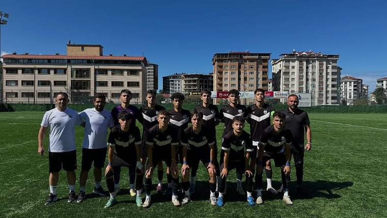 Futbol sahasında siyah formalı genç oyuncular ve iki kişi, şehir binaları önünde takım halinde duruyor.