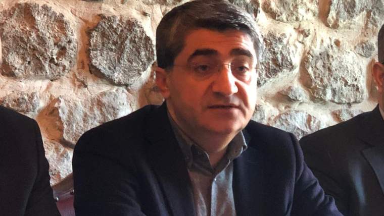Taş duvar önünde, koyu ceketli ve gözlüklü bir kişi masada otururken yanlarında iki kişi görülüyor.