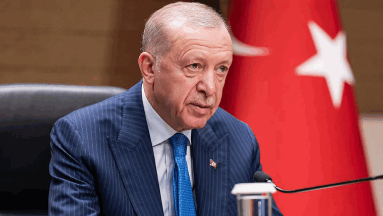 Mikrofon önünde oturan takım elbiseli bir kişi, arka planda Türk bayrağıyla görülüyor.