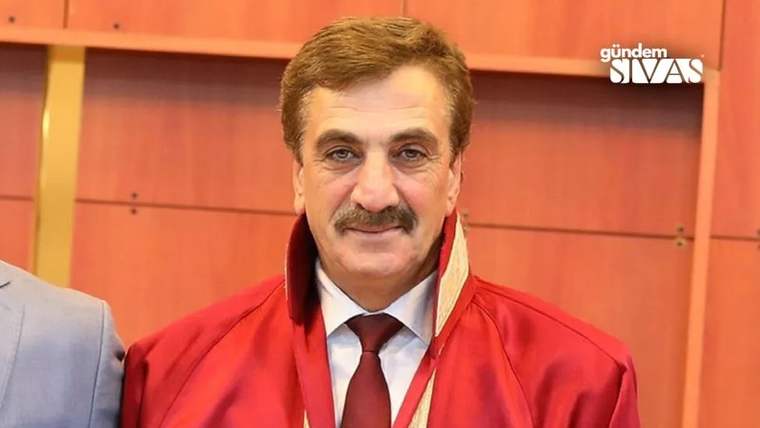 Kırmızı bir ceket giymiş, gülümseyen bir adam, arka planda ahşap bir duvar var.