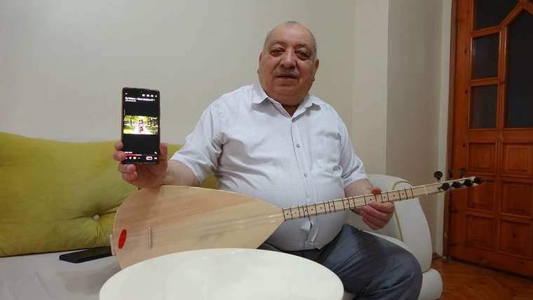 Bir adam, elinde bir akıllı telefon ve bir bağlama tutuyor. Telefon ekranında bir video açık.
