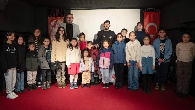 Kırmızı halı üzerinde, perde önünde yan yana duran çocuklar ve üç yetişkin görülüyor.