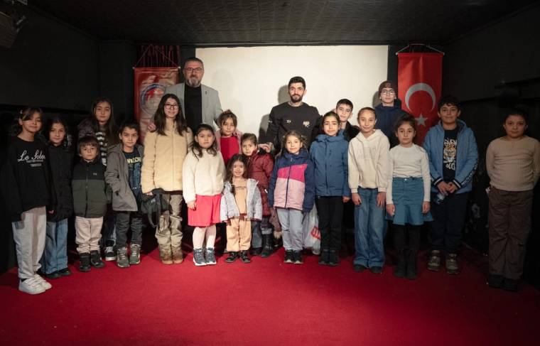 Kırmızı halı üzerinde, perde önünde yan yana duran çocuklar ve üç yetişkin görülüyor.