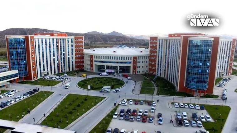 Kırmızı ve beyaz cepheli geniş hastane kompleksi, önündeki yollar, otopark ve dağ manzarasıyla görülüyor.