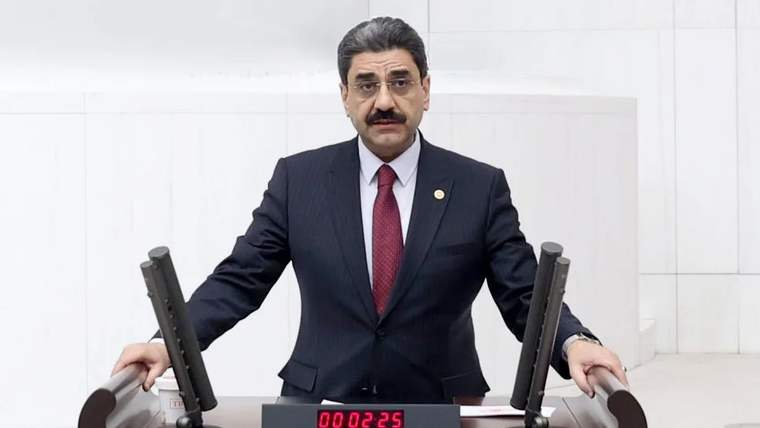 Kürsüde konuşan bir kişi, arkasında beyaz duvar ve saat var.