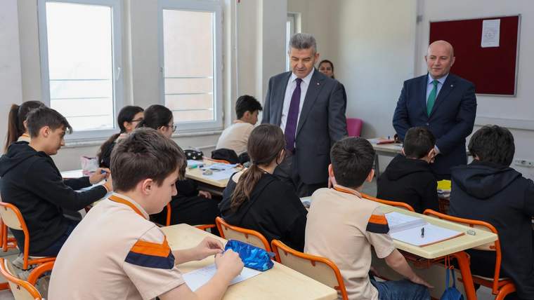 Sınıfta sıralarında oturan öğrencilerin yanında iki takım elbiseli kişi duruyor.