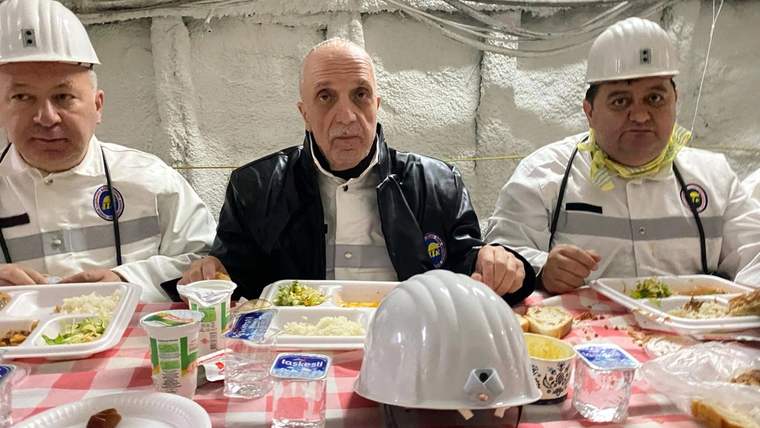 İftar sofrasında oturan üç kişi, önlerinde yemek tabakları var. Herkes beyaz kask takıyor.