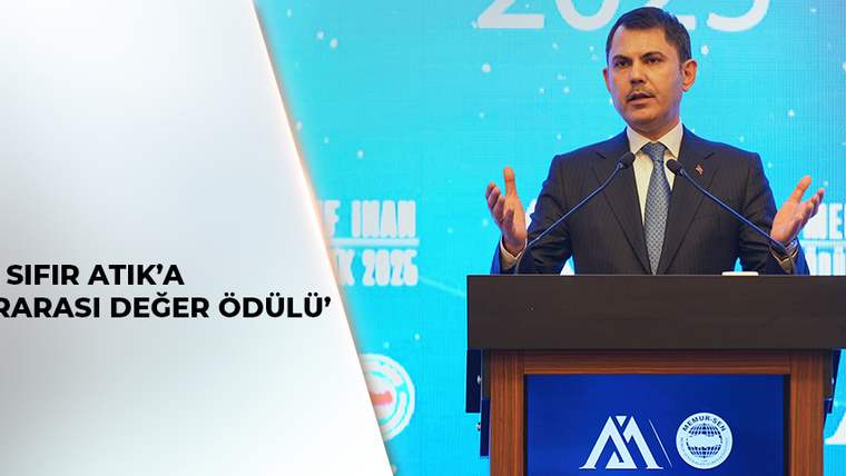 Takım elbiseli bir kişi, iki mikrofonlu kürsüde konuşurken arka planda mavi etkinlik ekranı görülüyor.