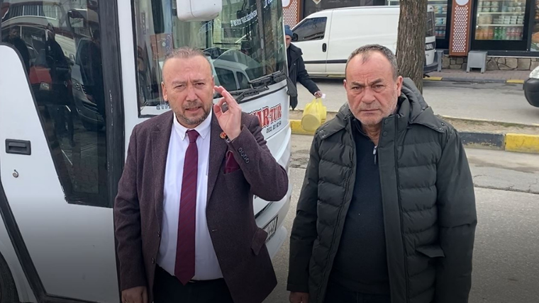 Bir otobüsün önünde duran iki erkekten biri elini kulağına götürürken diğeri kameraya bakıyor.