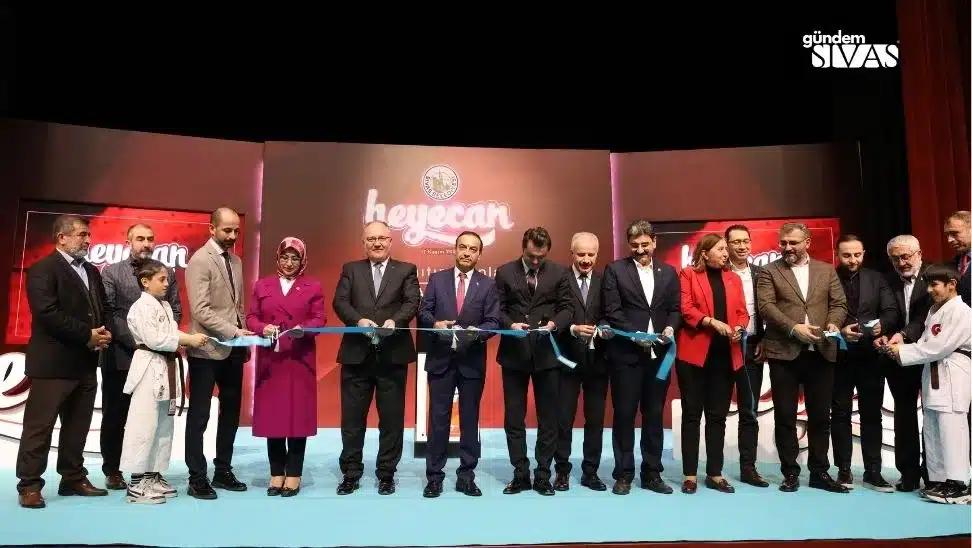 Sahnede bir grup yetişkin ve çocuk, mavi kurdele tutarak yan yana duruyor; arkada “heyecan” yazısı görülüyor.