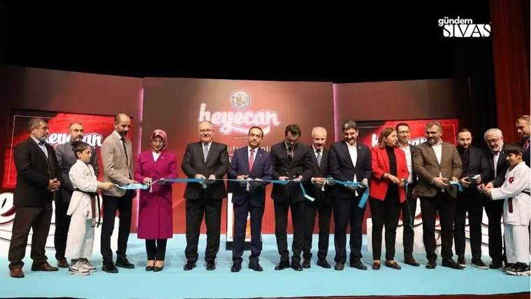Sahnede bir grup yetişkin ve çocuk, mavi kurdele tutarak yan yana duruyor; arkada “heyecan” yazısı görülüyor.