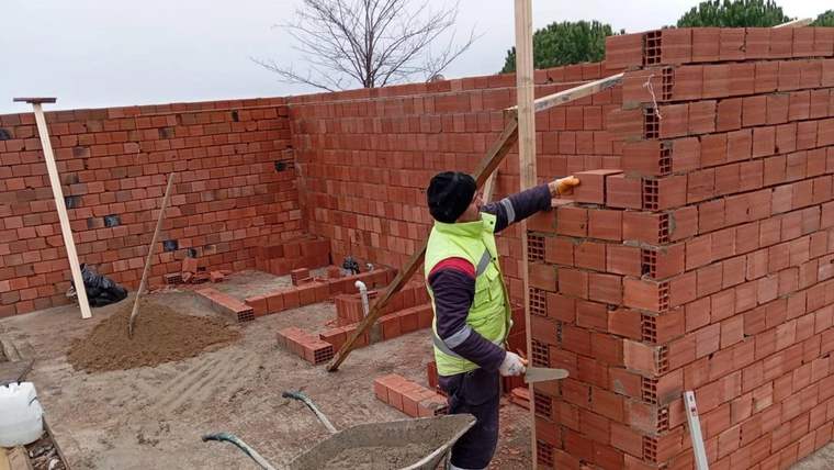 Yeşil yelekli bir işçi, tuğla duvar örülen alanda elindeki aletle duvarı işaret ediyor.