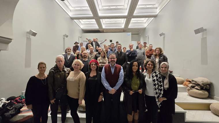 Aydınlık bir salonda, farklı yaşlardan yaklaşık 30 kişi birlikte durarak kameraya bakıyor.