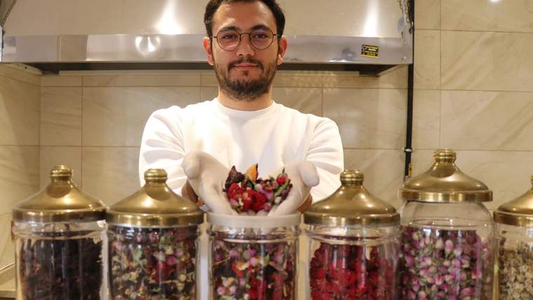 Mutfakta beyaz kıyafetli bir kişi, renkli kurutulmuş bitkilerle dolu cam kavanozların arkasında duruyor.