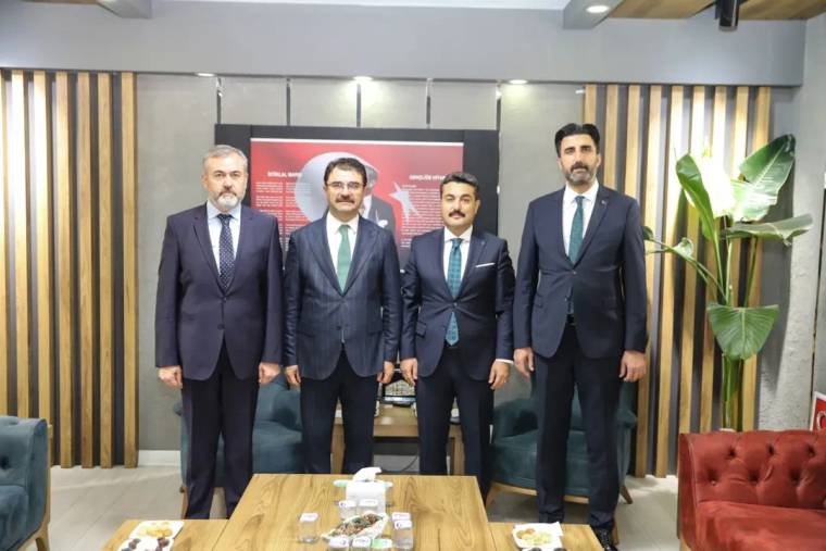 İç mekânda takım elbiseli dört kişi, arkalarında kırmızı panolu duvar önünde yan yana duruyor.