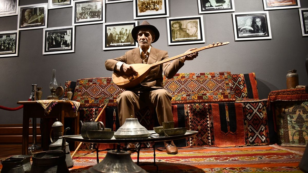 Duvarında çerçeveli fotoğraflar bulunan salonda, saz çalan bir kişi halılarla kaplı sedirde oturuyor.