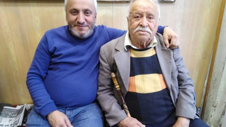Berber dükkânında yan yana oturan, biri mavi kazaklı iki erkek; yaşlı adam baston tutuyor.
