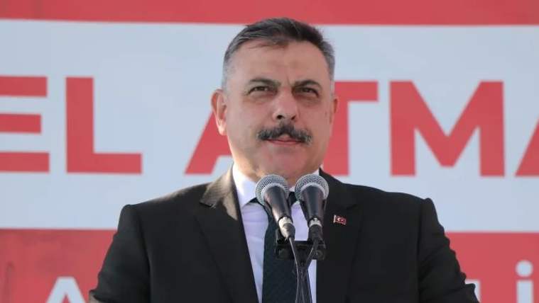 Kürsüde mikrofon önünde konuşan bir kişi, arka planda büyük bir pankart yer alıyor.