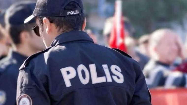 Sırtında ve şapkasında “POLİS” yazan üniformalı bir görevli, kalabalık önünde duruyor.