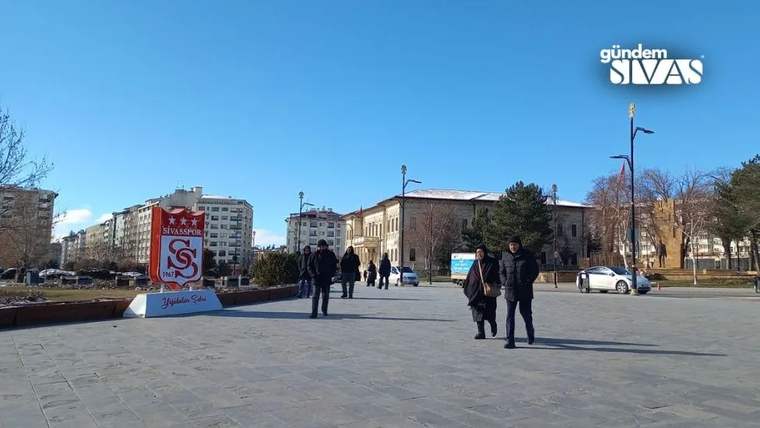 Açık havada, geniş taş meydanda yürüyen insanlar ve üzerinde Sivas arması bulunan pano görülüyor.