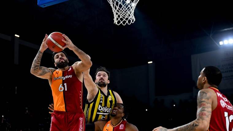 Basketbol maçında Galatasaray forması giyen bir oyuncu pota altında şut atarken rakipleri savunma yapıyor.