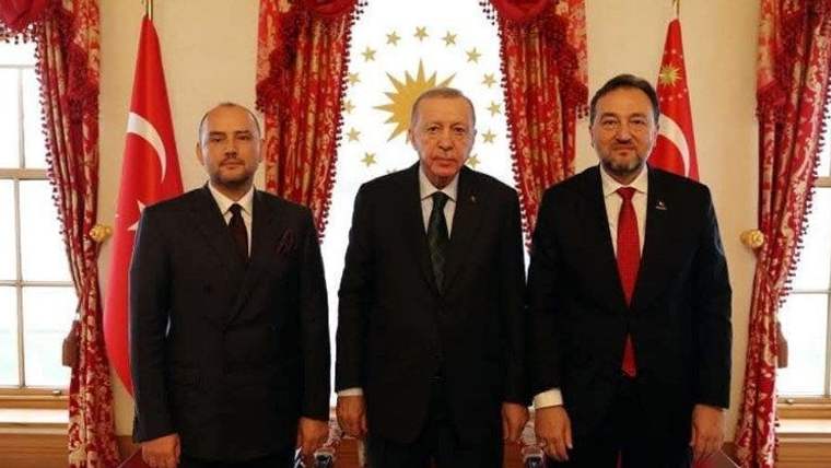 Üç kişi, arka planda Türk bayrakları ve bir sembol ile birlikte poz veriyor.