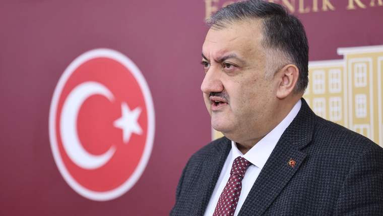 Takım elbiseli ve kravatlı bir kişi, kırmızı arka plan önünde kürsüde konuşurken görülüyor.