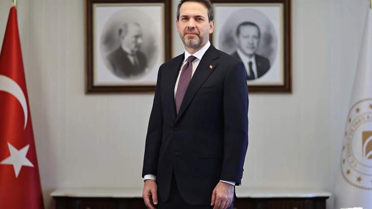 Resmi bir kıyafet giymiş bir kişi, arkasında iki çerçeveli portre ve Türk bayrağı ile duruyor.