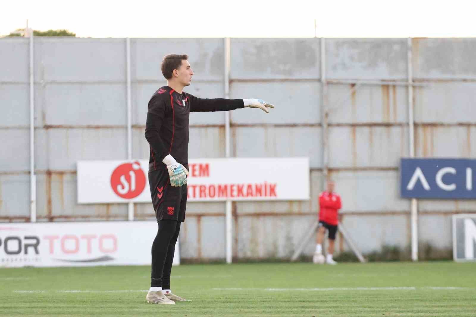 Siyah antrenman kıyafetli bir kaleci, futbol sahasında sağa doğru eliyle işaret ediyor.