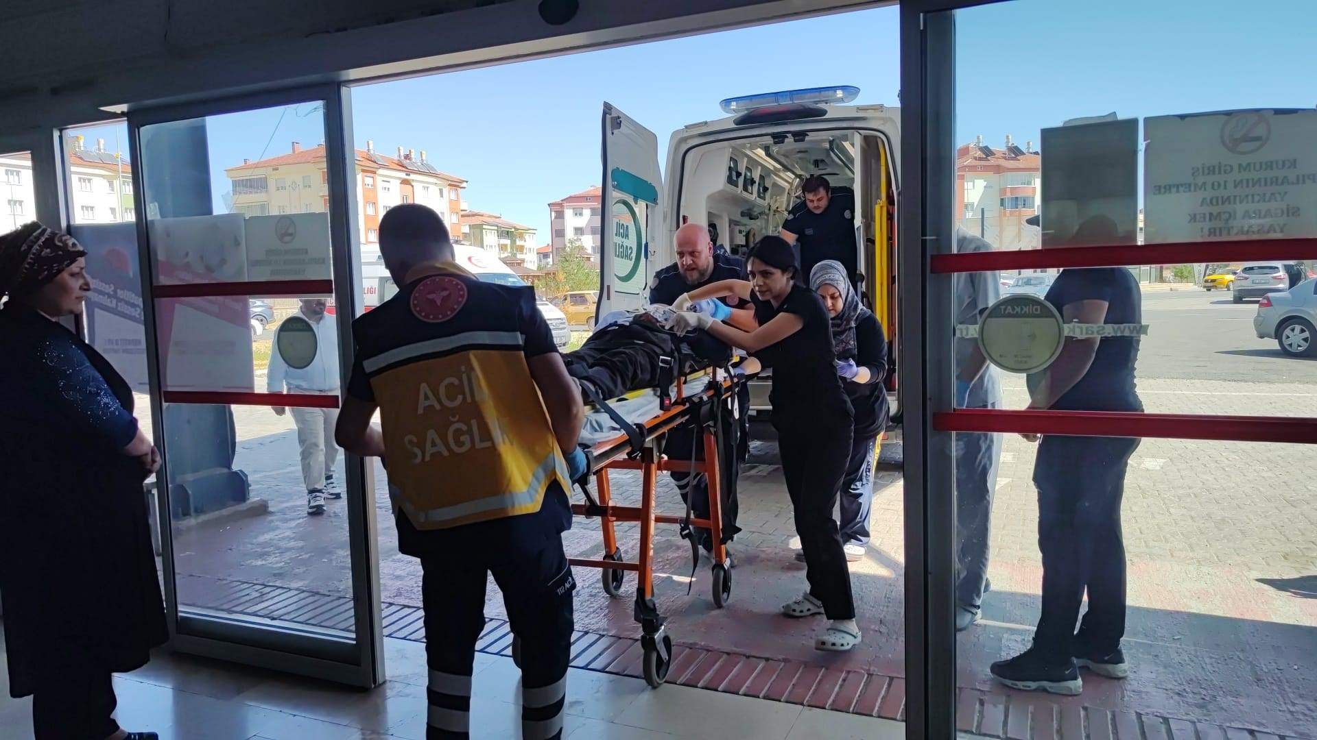 Ambulansın önünde, sedye üzerinde bir kişi taşınıyor; sağlık ekipleri ve diğer kişiler yardım ediyor.