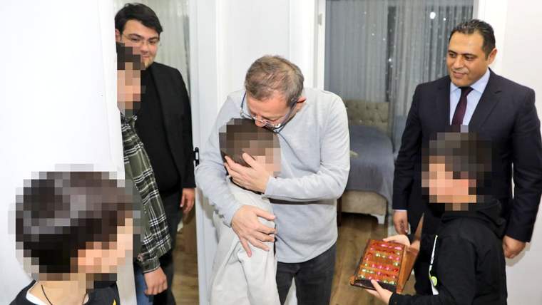 Bir yetişkin, çocukla sarılırken yanında takım elbiseli bir kişi ve elinde ikram tepsisi tutan çocuk görülüyor.