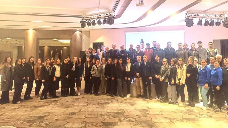 Toplantı salonunda, arka plandaki perde önünde çok sayıda kişinin birlikte verdiği grup pozu