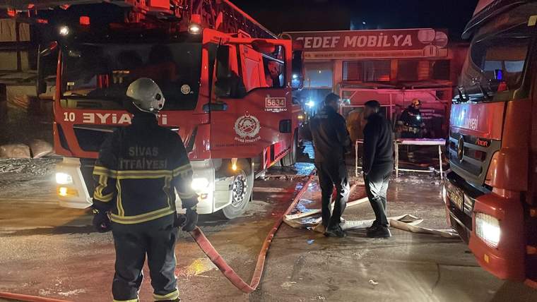 Gece vakti itfaiye araçları önünde hortum tutan itfaiyeci ve su birikintisi yanında iki kişi görülüyor.