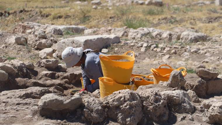 Taşlık bir alanda şapkalı bir kişi, turuncu kovalar ve kazı araçları arasında çalışıyor.